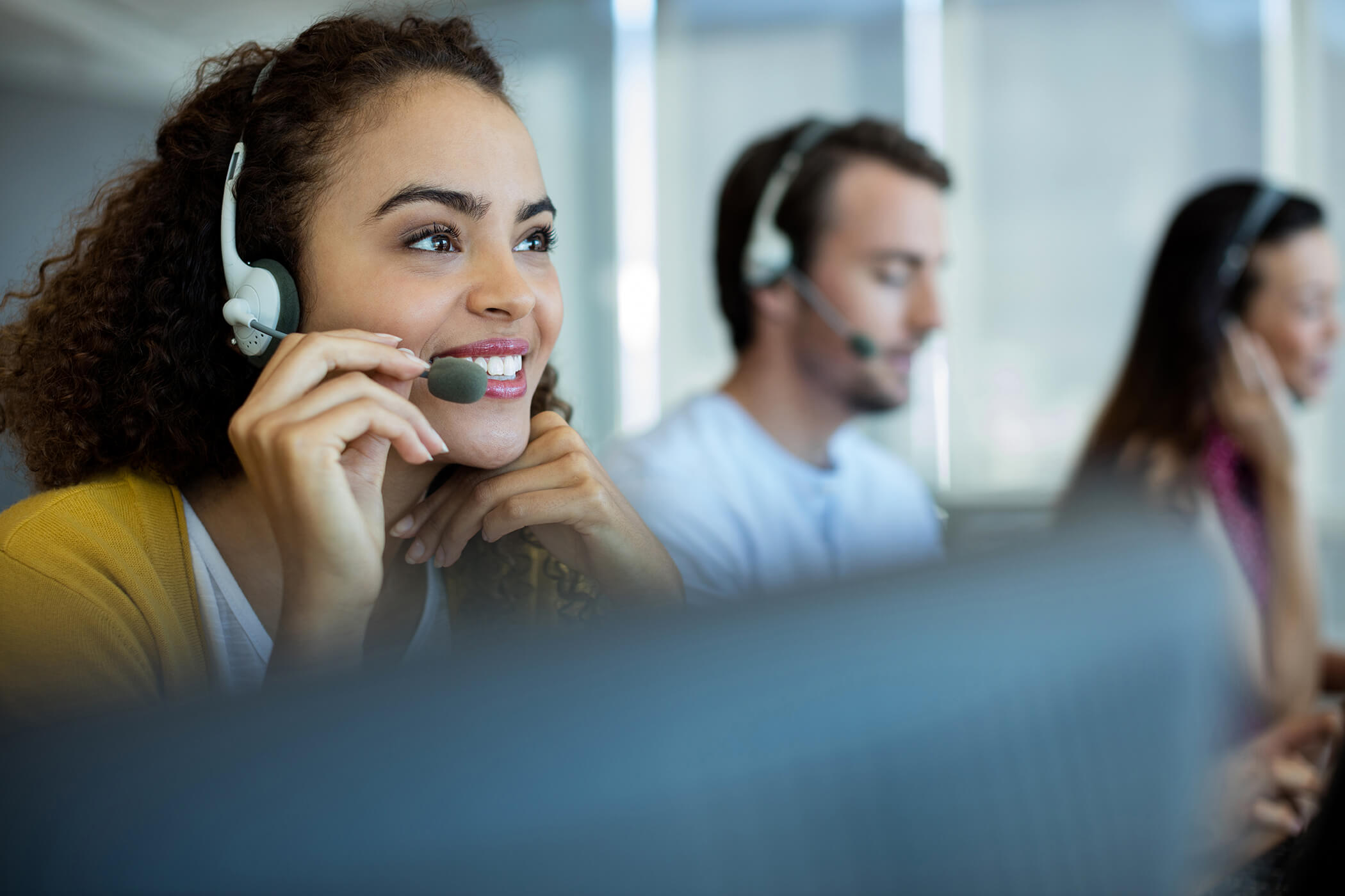  A smiling customer service representative wearing a headset is talking on the phone while two other customer service representatives work in the background.